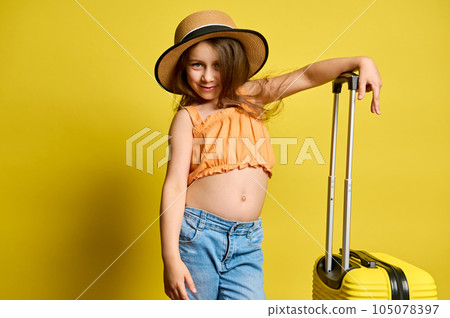 Happy child girl with stylish suitcase, cutely smiles at camera over yellow background. Travel, kids and summer concept 105078397