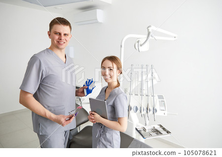 Two professional confident dentists posing near dental chair at clinic. Dentist and assistant in exam room smiling 105078621