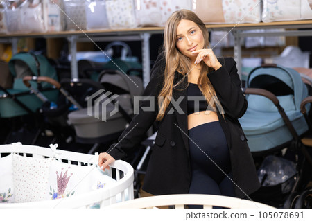 Pregnancy shopping concept. Pregnant woman choosing cradle cot or crib for newborn at baby shop store Pregnancy shopping concept. Pregnant woman choosing cradle cot or crib for newborn at baby shop store 105078691