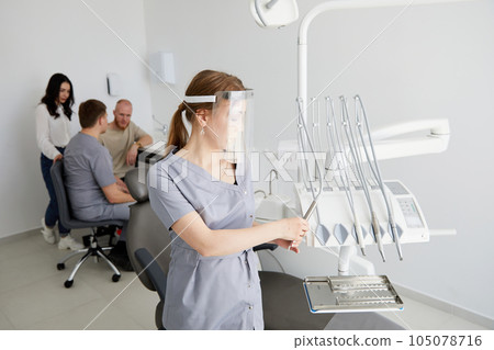 A young pretty female dentist stands in the middle of the office, and in the background a colleague is advising patients. A young pretty female dentist stands in the middle of the office, and in the background a colleague is advising patients. 105078716
