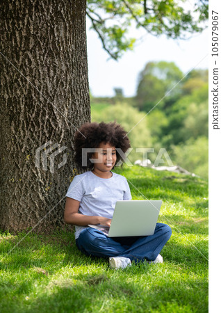 Little girl with afro hairstyle using laptop at park while sitting under tree. 105079067