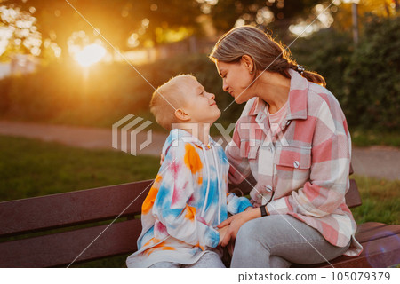 mother and son sit on a park bench in the rays of the setting sun. the concept of a family. Mother's Day. beautiful girl (mother) with a boy (son) in the park in the park are sitting on a bench at mother and son sit on a park bench in the rays of the setting sun. the concept of a family. Mother's Day. beautiful girl (mother) with a boy (son) in the park in the park are sitting on a bench at 105079379