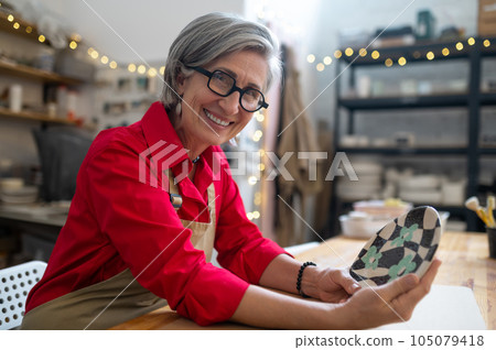 Woman potter in apron posing in ceramic workshop, holding crafted plate, looking at camera. Woman potter in apron posing in ceramic workshop, holding crafted plate, looking at camera. 105079418