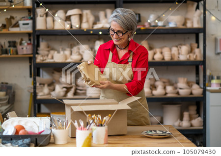 Young seller or worker of earthenware shop packing order of client and wrapping it into paper while standing by table against shelves 105079430