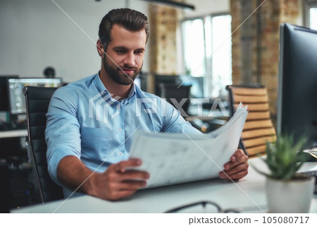 Great work Concentrated bearded engineer in formal wear looking at blueprint and smiling while sitting in the modern office 105080717