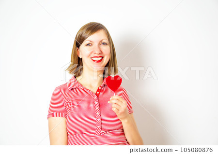 Beautiful woman in red and white shirt on a white background with a red heart lollipop in her hand. Beautiful woman in red and white shirt on a white background with a red heart lollipop in her hand. 105080876