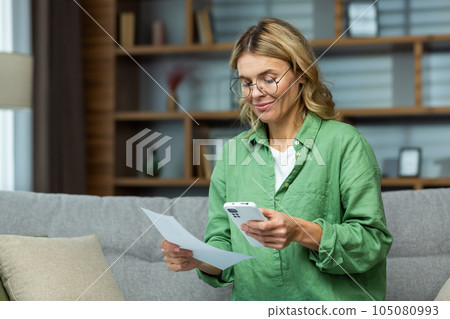 Mature experienced woman sitting on sofa at home, using app on smartphone to pay credit debts, smiling with satisfaction at ease and convenience of use. Mature experienced woman sitting on sofa at home, using app on smartphone to pay credit debts, smiling with satisfaction at ease and convenience of use. 105080993