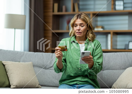 Cheated sad woman trying to make online payment fraudulently sitting on sofa in living room depressed holding bank credit card and smartphone. 105080995