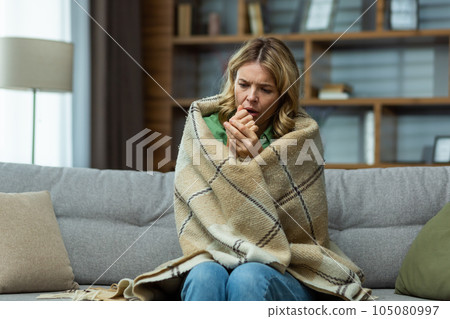 Single mature woman sitting at home in the living room on the sofa, wrapped in a blanket trying to keep warm, missing and broken heating in the house. Single mature woman sitting at home in the living room on the sofa, wrapped in a blanket trying to keep warm, missing and broken heating in the house. 105080997
