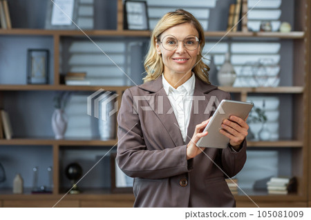 Mature experienced woman working inside office at workplace, portrait of businesswoman holding tablet computer, boss smiling and looking at camera. 105081009