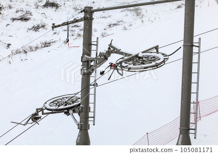 Large metal wheels for steel cables on top of ski lift supporting pillar, snow covered piste in background 105081171