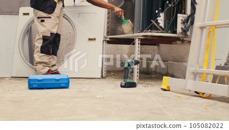 Ground level shot of trained engineer commissioned by home owner for yearly air conditioner routine maintenance, using soft dusting brush to clean dusty condenser coils and filters, outdoor 105082022