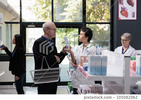 Medical store employee advising old client sunblock cream, talking with customer in apothecary aisle. Elderly buyer and young pharmaceutical assistant discussing sunscreen near medicaments shelf Medical store employee advising old client sunblock cream, talking with customer in apothecary aisle. Elderly buyer and young pharmaceutical assistant discussing sunscreen near medicaments shelf 105082062