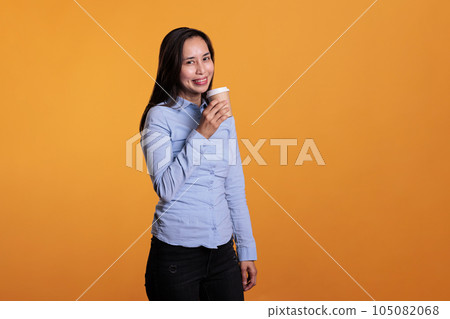 Asian woman holding cup of coffee drinking hot beverage enjoying work break before start to shoot in studio. Excited fashionable model with brunette hair standing over yellow background Asian woman holding cup of coffee drinking hot beverage enjoying work break before start to shoot in studio. Excited fashionable model with brunette hair standing over yellow background 105082068