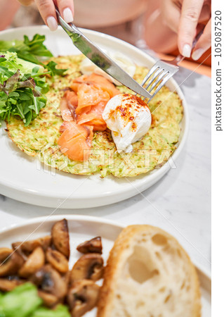 Woman cuts Zucchini pancakes, with avocado, cheese cream, salmon and egg . Healthy breakfast, protein. Restaurant dish. Breakfast in cafe on a sunny morning, on the summer veranda Woman cuts Zucchini pancakes, with avocado, cheese cream, salmon and egg . Healthy breakfast, protein. Restaurant dish. Breakfast in cafe on a sunny morning, on the summer veranda 105087520