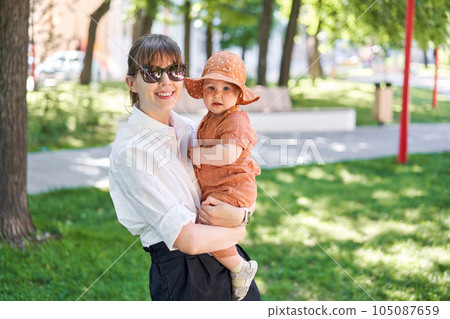 Portraits young mother and her daughter of acute 10 months old baby girl. Walking on the playground, on a sunny summer day. 105087659