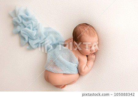 A cute newborn boy in the first days of life sleeps naked against a background of white fabric. Blue light cape, transparent scarf. A cute newborn boy in the first days of life sleeps naked against a background of white fabric. Blue light cape, transparent scarf. 105088342