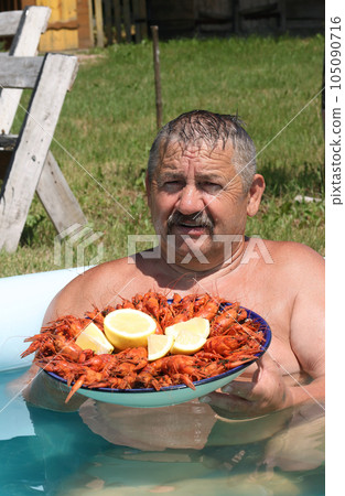 Joyful mustachioed man sits in the pool. He holds a large dish with boiled crayfish in his hands. Crayfish are decorated with lemon slices. Close-up. Joyful mustachioed man sits in the pool. He holds a large dish with boiled crayfish in his hands. Crayfish are decorated with lemon slices. Close-up. 105090716