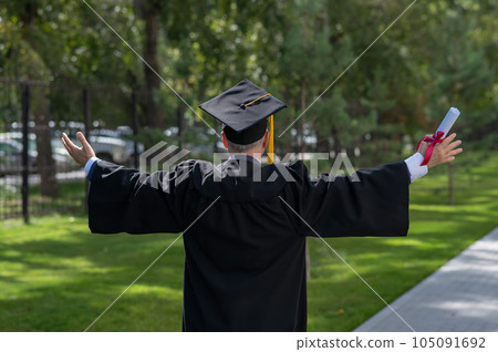 Elderly male graduate rejoices at graduation outdoors. Rear view of a man in a robe with arms outstretched to the sides. Elderly male graduate rejoices at graduation outdoors. Rear view of a man in a robe with arms outstretched to the sides. 105091692
