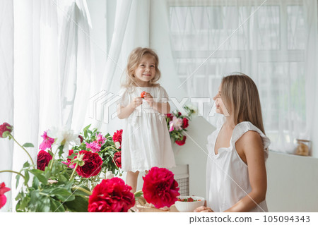 A little blonde girl with her mom on a kitchen countertop decorated with peonies. The concept of the relationship between mother and daughter. Spring atmosphere. A little blonde girl with her mom on a kitchen countertop decorated with peonies. The concept of the relationship between mother and daughter. Spring atmosphere. 105094343