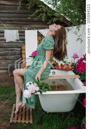 A woman is sitting on a cast-iron bathtub in the courtyard of a country house next to a bush of flowering peonies. The concept of summer, country life, a bathroom on the street in a blooming garden in A woman is sitting on a cast-iron bathtub in the courtyard of a country house next to a bush of flowering peonies. The concept of summer, country life, a bathroom on the street in a blooming garden in 105094523