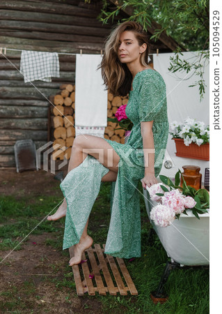 A woman is sitting on a cast-iron bathtub in the courtyard of a country house next to a bush of flowering peonies. The concept of summer, country life, a bathroom on the street in a blooming garden in 105094529