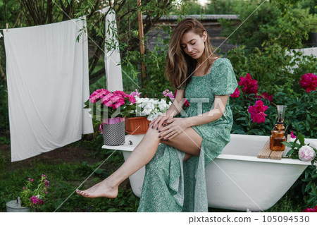 A woman is sitting on a cast-iron bathtub in the courtyard of a country house next to a bush of flowering peonies. The concept of summer, country life. 105094530
