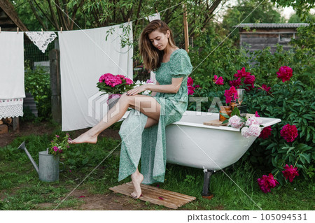 A woman is sitting on a cast-iron bathtub in the courtyard of a country house next to a bush of flowering peonies. The concept of summer, country life. 105094531