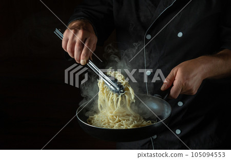 The cook fries spaghetti in a hot pan. Space for advertising on a black background. Italian national cuisine The cook fries spaghetti in a hot pan. Space for advertising on a black background. Italian national cuisine 105094553