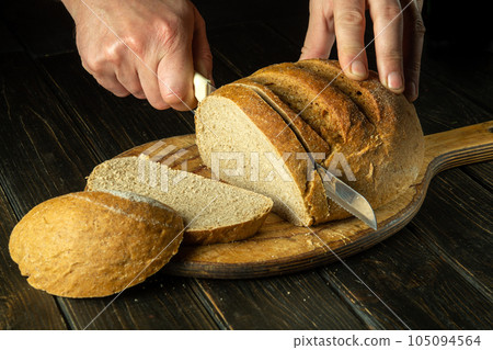 The chef hand with a knife slices bread on a kitchen cutting board. The concept of healthy food in the bakery. The chef hand with a knife slices bread on a kitchen cutting board. The concept of healthy food in the bakery. 105094564