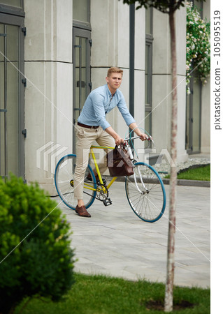 Handsome, elegant businessman in smart casual clothes going to office on bike, riding along city with modern buildings 105095519