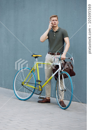 Handsome, concentrated, stylish businessman in smart casual clothes standing outdoors with bike and talking on mobile phone 105095569