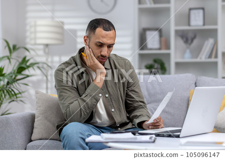 Tired and thoughtful african american man working from home on laptop with documents and bills. Overtime over the deadline. Tired and thoughtful african american man working from home on laptop with documents and bills. Overtime over the deadline. 105096147