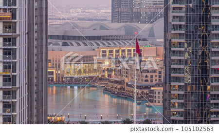 Aerial view of Dubai Fountain in downtown with palms in park next to shopping mall and souq day to night timelapse, UAE Aerial view of Dubai Fountain in downtown with palms in park next to shopping mall and souq day to night timelapse, UAE 105102563