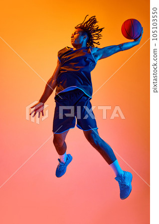 Full-length image of young sportsman, basketball player in motion, jumping with ball against orange background in neon lights 105103550