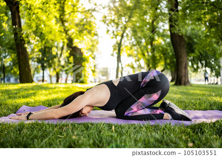 Girl doing morning yoga staying in child pose at park outdoors in summer time. Young woman exercising at nature in sunny day 105103551