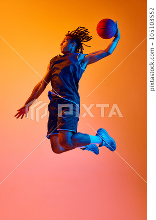 Full-length image of young sportsman, basketball player in motion, jumping with ball against orange background in neon lights Full-length image of young sportsman, basketball player in motion, jumping with ball against orange background in neon lights 105103552