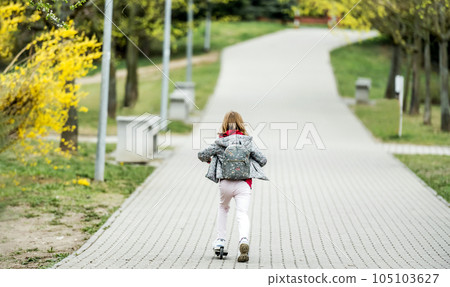 Little cute girl riding a scooter on a path in the park, back view Little cute girl riding a scooter on a path in the park, back view 105103627
