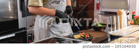 Chef in uniform warming up ready dish with fire on restaurant kitchen before serving to client 105104030