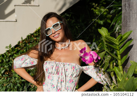 Young beautiful woman in a romantic dress with a floral print, sunglasses and a pearl necklace bracelet, against the backdrop of tropical leaves Young beautiful woman in a romantic dress with a floral print, sunglasses and a pearl necklace bracelet, against the backdrop of tropical leaves 105104112
