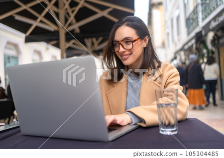 Female student, freelancer in glasses using laptop computer sitting at table in outdoor cafe 105104485