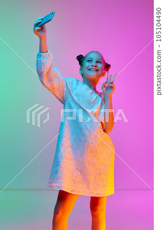 Happy school-age girl, child wearing white dress using cell phone over neon light background. Concept of childhood, emotions, kids fashion 105104490