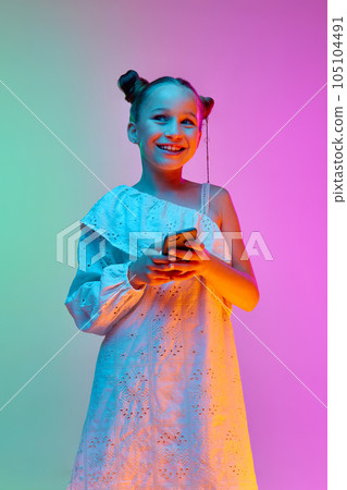 Happy school-age girl, child wearing white dress using cell phone over neon light background. Concept of childhood, emotions, kids fashion Happy school-age girl, child wearing white dress using cell phone over neon light background. Concept of childhood, emotions, kids fashion 105104491