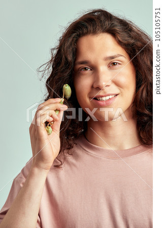 Young handsome long haired guy using jade de-puffing face roller to keep his skin soft and smooth. Caucasian man with brown hair maintaining his natural beauty with skincare routine. Men's self-care. 105105751