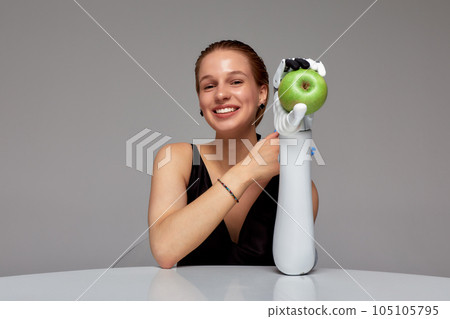 Young smiling girl with disability wearing sensory bionic prosthetic arm seats at the table. Beautiful woman holds a green apple in her raised artificial robotic hand. Full life after limb loss. Young smiling girl with disability wearing sensory bionic prosthetic arm seats at the table. Beautiful woman holds a green apple in her raised artificial robotic hand. Full life after limb loss. 105105795