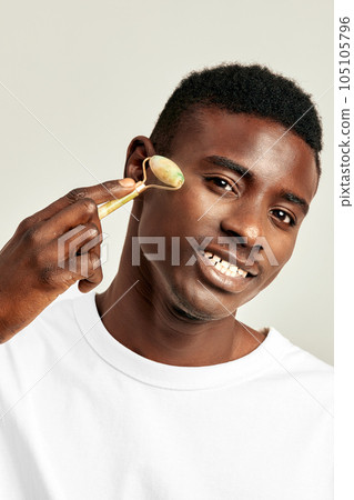 Young handsome black guy using jade de-puffing face roller to keep his skin soft and smooth. African American millennial man maintains his natural beauty with a skincare routine. Men's self-care. Young handsome black guy using jade de-puffing face roller to keep his skin soft and smooth. African American millennial man maintains his natural beauty with a skincare routine. Men's self-care. 105105796