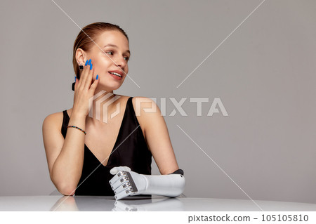 Young girl with disability wearing sensory bionic prosthetic arm seats at the table in studio. Beautiful woman freely controls artificial robotic hand . Full life after limb loss. 105105810