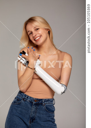 Studio half-length portrait of beautiful young blonde girl with disability wearing sensory bionic prosthetic arm. Woman dressed in blue jeans and beige top freely controls artificial robotic hand. 105105869
