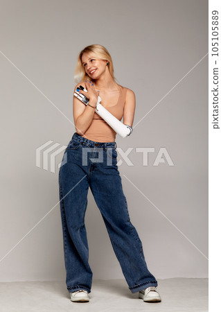 Studio portrait of beautiful young blonde girl with disability wearing sensory bionic prosthetic arm. Woman dressed in blue jeans and beige top posing and freely controls artificial robotic hand. 105105889
