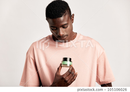 Young handsome black guy holds a jar of moisturizing healing skin creme in his hand and going to use it. African American millennial man practices skincare routine to keep healthy and youthful look. 105106026